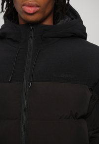 Gros plan sur une personne portant une doudoune noire à capuche avec une fermeture éclair centrale et un logo discret "Han Kjøbenhavn" sur la poitrine.