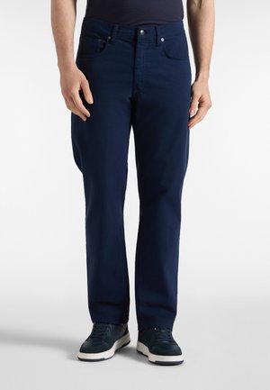 Uomo che indossa jeans blu scuro e scarpe da ginnastica nere, in piedi davanti a uno sfondo bianco uniforme.