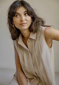 Femme aux cheveux ondulés mi-longs, vêtue d'une robe beige sans manches boutonnée, regardant pensivement sur le côté sur un fond uni.