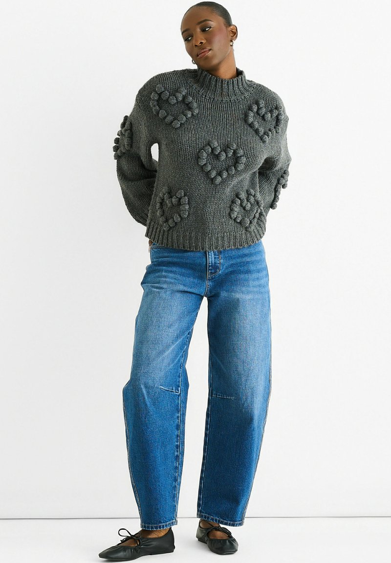 Gray knitted sweater with heart-shaped bobble details, paired with loose blue jeans and black flat shoes. Round neckline and textured fabric.