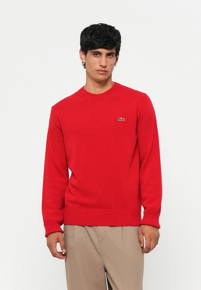 Pulover tricotat roșu cu guler rotund, mansete și tiv cu brâu. Prezintă un mic logo verde Lacoste pe partea stângă a pieptului.