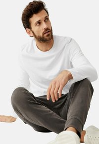 Weißes Langarmhemd kombiniert mit grauen Hosen, sitzend. Das Hemd hat einen Rundhalsausschnitt; die Hose hat ein tailliertes Design und elastische Bündchen. Weiße Sneaker.