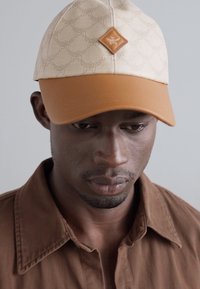 Casquette crème avec tissu à motifs, dessous beige et visière brune. Présente un écusson de logo brun à l'avant. Chemise brune partiellement visible.
