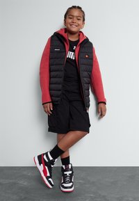 Gilet imbottito nero con maniche rosse, pantaloni corti neri e sneakers alte nere, rosse e bianche. Maglietta scura con logo bianco. Posizione sorridente.