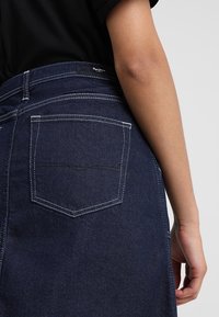 Falda de mezclilla azul oscura con costuras blancas. Presenta un bolsillo trasero y una cinturilla visible, mostrando un diseño clásico y un corte regular.