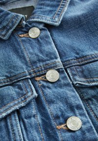 Chaqueta de mezclilla con un lavado azul claro, cuenta con botones a presión metálicos plateados, bolsillos con solapa y costuras en contraste, mostrando un acabado texturizado.