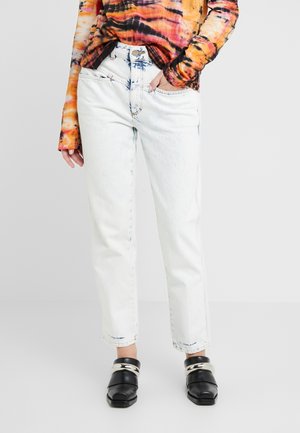 Person iført lystvaskede straight-leg jeans, sorte sko med firkantet tå og sølvkæde-detalje samt en langærmet orange og sort tie-dye top.