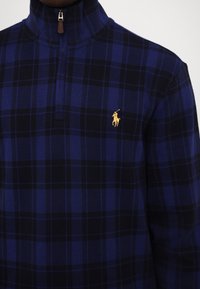 Pull à motif à carreaux bleu et noir avec une fermeture éclair quart de longueur et un logo brodé jaune représentant un joueur de polo sur la poitrine.