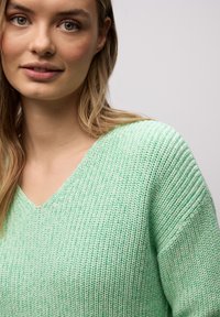 Jeune femme aux longs cheveux blonds portant un pull vert clair en maille à col en V, légèrement tournée vers la gauche sur un fond uni clair.