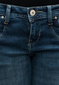 Κοντινό πλάνο της μέσης σκούρου μπλε τζιν παντελονιού με κουμπί που γράφει "Genuine Denim Goods" και εμφανείς λεπτομέρειες ραφής.