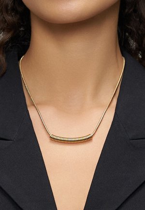 Goldkette mit einer gebogenen Stange mit Musterverzierung, die an einer dünnen Kette hängt und über einem schwarzen Kleidungsstück getragen wird.