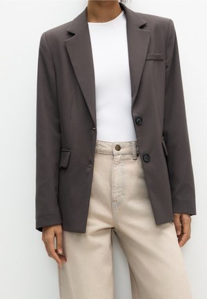 Personne portant un blazer gris foncé à double boutonnage sur un haut blanc et un pantalon beige taille haute, debout devant un fond blanc uni.