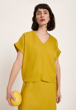 Woman with short dark hair wearing mustard yellow V-neck top and skirt, holding a yellow melon in her left hand.