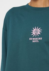 Зелен суитшърт с графика на розова спирала слънце над текста "SUNSHINE SOUL" на гърдите.
