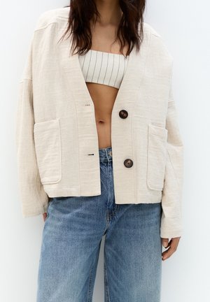 Femme portant une veste beige boutonnée par-dessus un crop top rayé avec un jean bleu taille haute, sur un fond clair uni.