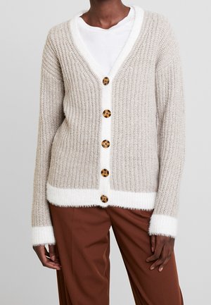 Nahaufnahme einer Person, die eine beige Strickjacke mit weißem Rand und braunen Knöpfen trägt, kombiniert mit rostfarbenen Hosen.