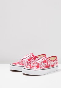 Rote Leinen-Sneaker mit einem weißen Grafikdruck und wiederholtem Text. Weiße Schnürsenkel, Gummisohle und schwarzer Akzent am Rand.