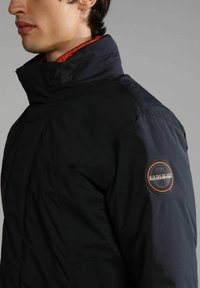 Giacca marina con colletto alto, fodera interna arancione e una patch circolare con logo sulla manica sinistra. Texture liscia e dettagli di taglio su misura.