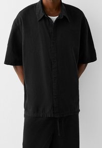 Muž nosící volnou černou košili s krátkým rukávem přes bílý podvlékací tričko, v kombinaci s černými kalhotami na jednobarevném pozadí.