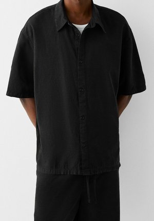 Hombre vistiendo una camisa negra de manga corta y holgada sobre una camiseta blanca, combinada con pantalones negros, frente a un fondo simple.