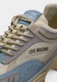 Αθλητικό παπούτσι Love Moschino σε μπεζ και μπλε χρώμα με υφασμάτινες και σουέντ υφές, μπεζ κορδόνια και λογότυπο της μάρκας στη γλώσσα και στην πλαϊνή πλευρά.