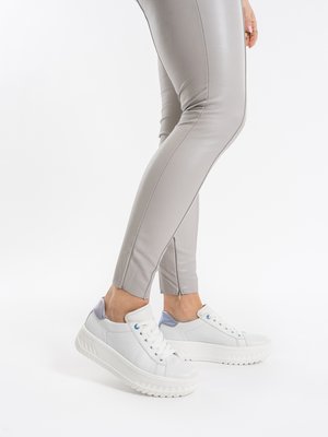 Person trägt hellgraue, eng anliegende Hose und weiße Plateau-Sneaker, steht mit einem leicht angehobenen Fuß vor weißem Hintergrund.