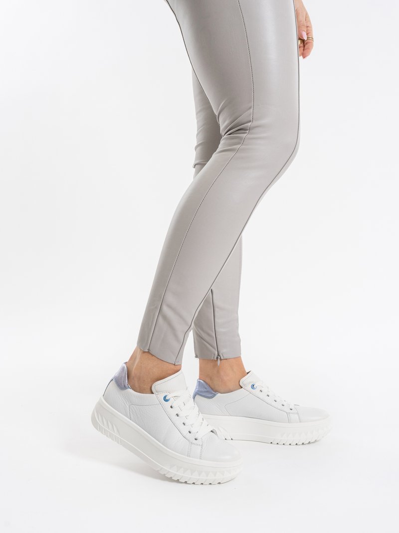 Person iført lysegrå, tætsiddende bukser og hvide platform-sneakers, stående med den ene fod let løftet på en hvid baggrund.