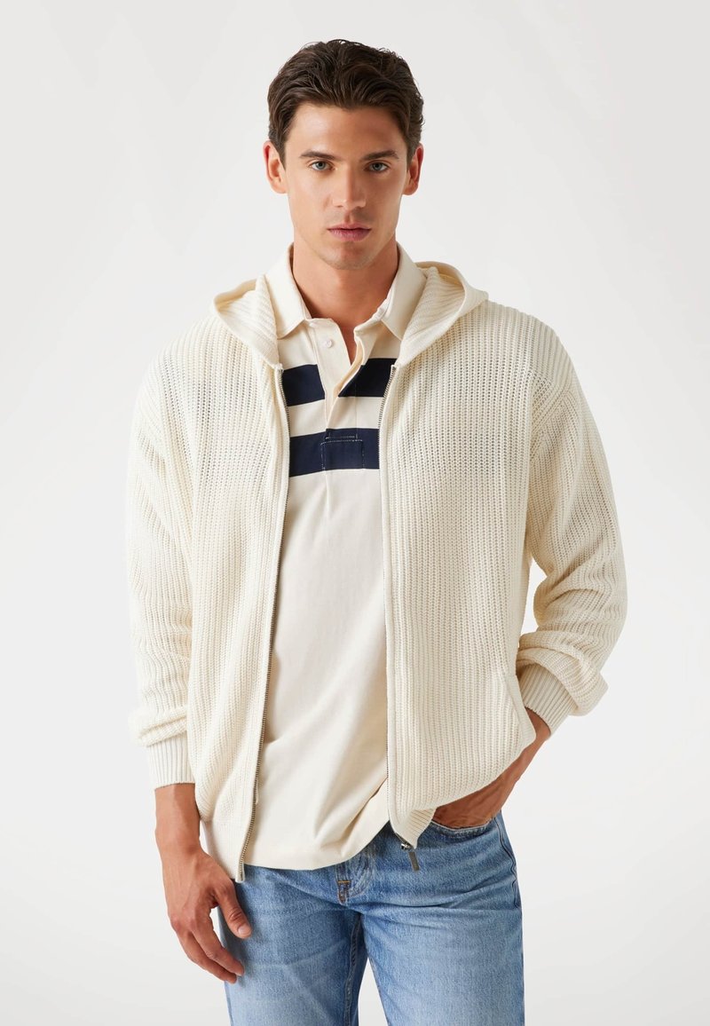 Hellbeige Zip-Up-Pullover mit strukturiertem Strickmuster und Kapuze, über einem cremefarbenen Poloshirt mit marineblauen Streifen getragen. Kombiniert mit hellblauen Jeans.