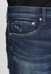 Granatowe dżinsy mają klasyczny design, widoczne przeszycia, małą przednią kieszeń i przetarcia na tkaninie.