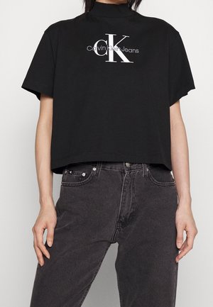 Osoba ubrana w czarny krótki t-shirt Calvin Klein Jeans i wyblakłe czarne jeansy z wysokim stanem, stoi z rękami swobodnie opuszczonymi wzdłuż ciała.