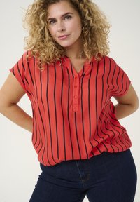Blusa roja con rayas verticales negras, mangas cortas, escote en V y cuatro botones a lo largo de la tapeta. Tejido suave y liso con ligera caída.