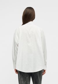 Personne aux longs cheveux bruns vue de dos, portant une chemise blanche ample à manches longues et un jean foncé délavé sur un fond uni.