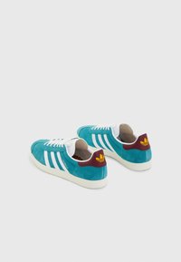 Baskets en daim turquoise avec trois bandes blanches, lacets blancs et un renfort de talon bordeaux avec un logo jaune. Tige texturée, semelle en caoutchouc.