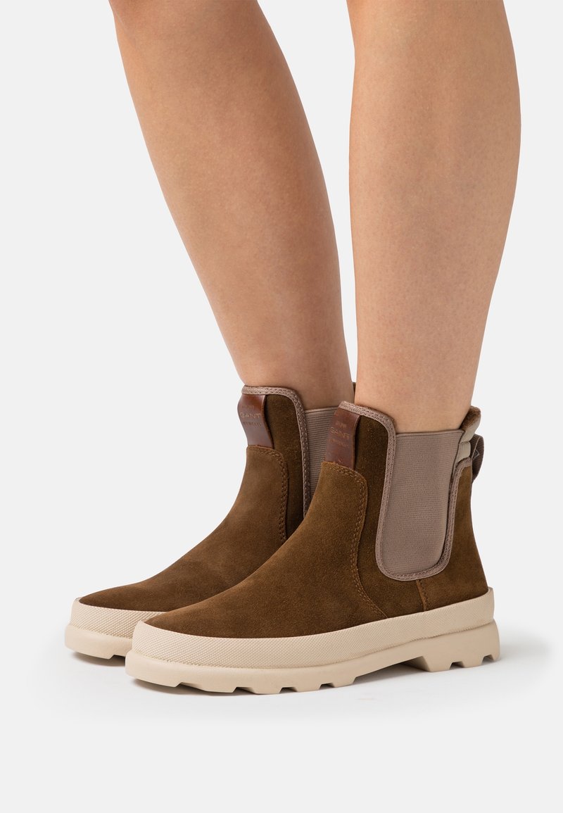 Bottines en daim marron avec panneaux latéraux élastiques beiges et semelles épaisses en caoutchouc, portées sur des jambes nues, présentées sur un fond blanc uni.