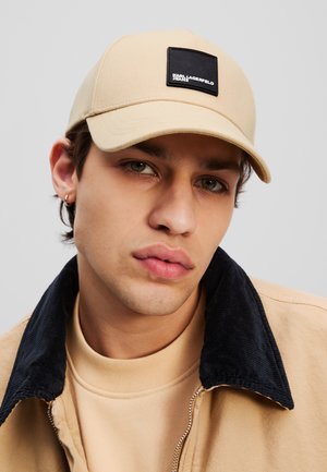 Gorra beige con visera redondeada y un parche cuadrado negro con texto blanco. Se lleva con un sudadera de color claro y un cuello de pana oscuro.