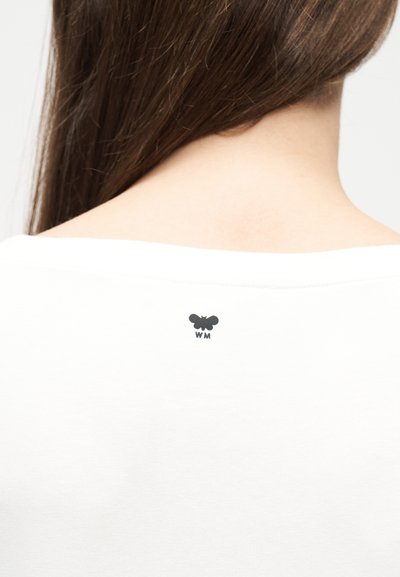 Πίσω όψη ενός ατόμου με μακριά καστανά μαλλιά που φοράει λευκό μπλουζάκι με ένα μικρό μαύρο λογότυπο πεταλούδας και τα γράμματα «WM» από κάτω στο άνω μέρος της πλάτης.