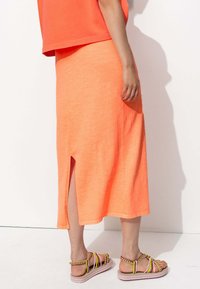 Jupe midi en coton orange avec fente latérale, associée à des sandales colorées à lanières avec une semelle épaisse. Design simple et décontracté.