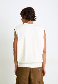 Ermeløs kremfarget strikkegenser med ribbestrikket nederkant, med en glatt tekstur og et enkelt design. Kombinert med brune cargo-shorts.