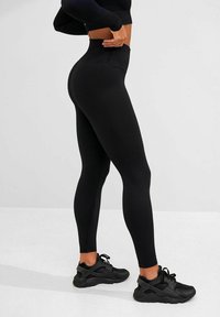 Kvinde iført sorte højtaljede leggings og sorte sportssko, stående sidelæns med hænderne på hoften mod en ensfarvet baggrund.