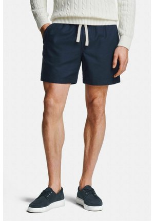 Man draagt marineblauwe shorts met trekkoord, een witte kabelgebreide trui en marineblauwe sneakers met witte zolen, staand met handen in de zakken.
