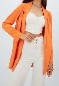Blazer arancione in tessuto leggero con vestibilità su misura. Indossato sopra un top corto in pizzo bianco, abbinato a pantaloni bianchi a vita alta.