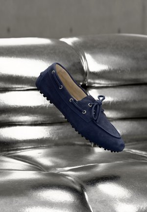 Mocassin en daim bleu marine avec lacets flottant au-dessus d'un canapé en cuir capitonné argenté métallique brillant.