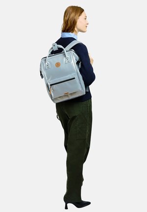 Frau trägt dunklen Pullover und grüne Hose, hat einen hellgrauen quadratischen Rucksack mit Fronttasche und oberen Tragegriffen.