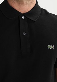 Camisa polo negra hecha de tejido texturizado, con cuello, dos botones y un pequeño logotipo de cocodrilo verde en el lado izquierdo del pecho.