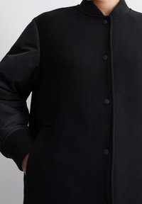 Giacca bomber nera con corpo in lana e maniche in tessuto lucido, polsini a costine, bottoni a pressione e tasche laterali. Design minimalista con una finitura testurizzata.