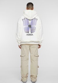 Hoodie blanc à coupe oversize, avec un grand motif de papillon violet dans le dos, associé à un pantalon cargo beige.