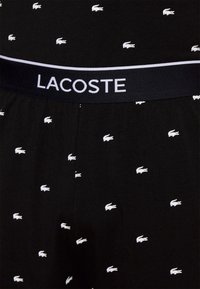 Sorte boxer shorts med en hvid Lacoste-logo i taljen og gentagne små hvide krokodillemønstre over stoffet. Blødt bomuldsmateriale.