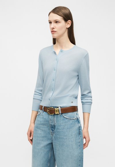 Vrouw draagt een lichtblauwe vest met knopen, lichtblauwe spijkerbroek en een bruine riem met een gouden gesp, staand tegen een egale achtergrond.