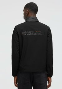 Chaqueta deportiva negra con cuello alto. Presenta frases impresas en diversas tipografías en la parte posterior. Textura suave, diseño entallado.