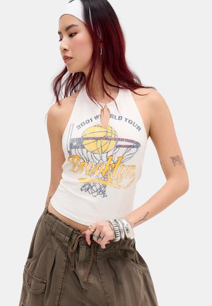 Giovane donna con capelli tinti di rosso che indossa un top halter bianco con stampa di pallacanestro e la scritta "Brooklyn", pantaloni cargo verde oliva e braccialetti d'argento.